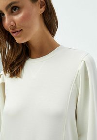 Sudadera beige clara con escote redondo, tejido texturizado y mangas abullonadas con detalles de costura sutiles.