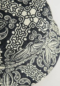 Cappello a secchiello a fantasia nera e crema con design floreali e geometrici, realizzato in tessuto testurizzato con una corona arrotondata e una visiera morbida.