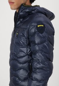 Giacca a vento blu navy con texture lucida, cappuccio regolabile e zip frontale. Presenta un logo a patch sulla spalla e due tasche per le mani.