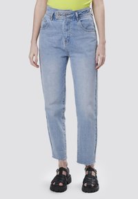 Jeans de talle alto en azul claro con corte recto, dobladillo deshilachado, dos bolsillos frontales y cierre de doble botón. Combinados con sandalias negras.