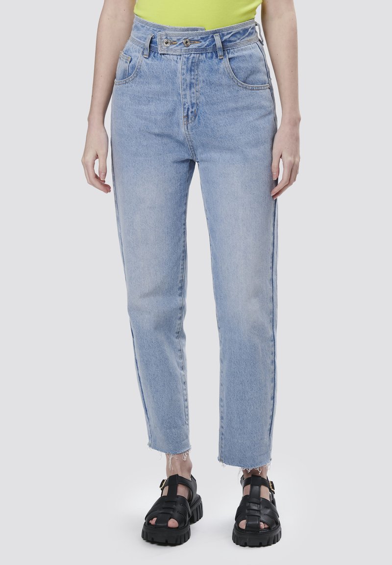 Jeans de talle alto en azul claro con corte recto, dobladillo deshilachado, dos bolsillos frontales y cierre de doble botón. Combinados con sandalias negras.