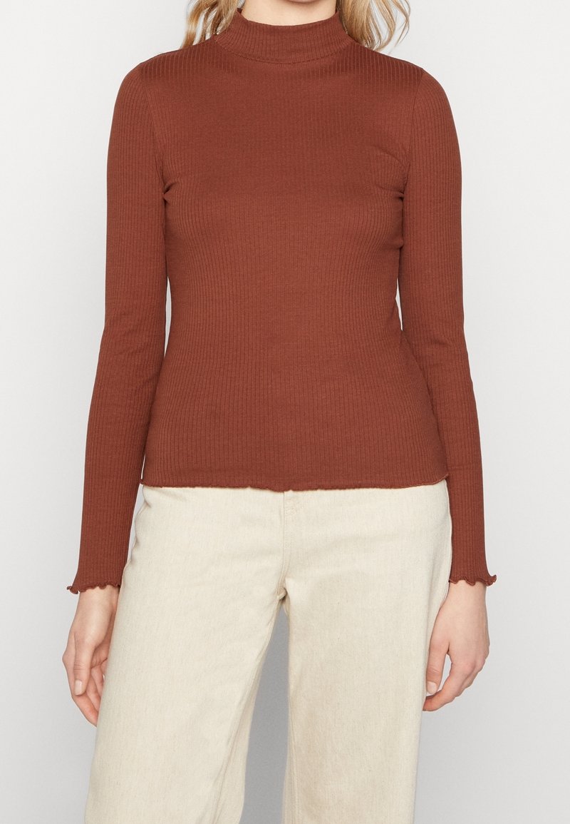 Braun gerippter Rollkragenpullover mit langen Ärmeln und gewellten Bündchen, aus einem weichen Stoff gefertigt, kombiniert mit hellen Hosen.