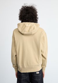 Pessoa com cabelo escuro e encaracolado vestindo um hoodie bege com o logotipo "Dickies" na capuz e calças azul-escuro, de costas para o espectador.
