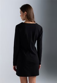 Robe noire à manches longues avec une silhouette droite, un col rond et des détails de couture dans le dos. Tissu lisse et coupe à hauteur des genoux.