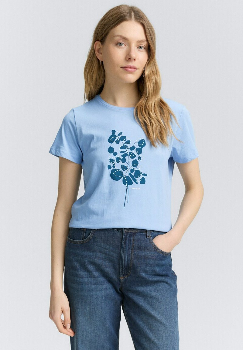 Camiseta de algodón azul claro con un gráfico floral en azul oscuro. Mangas cortas, escote redondo, corte holgado, combinada con jeans de mezclilla oscuros.