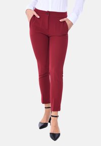 Pantaloni slim-fit bordeaux con una texture liscia, tasche frontali e chiusura a bottone, abbinati a décolleté nere a punta.