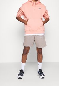 Ljusrosa huvtröja med framficka, matchad med beiga shorts med en liten logotyp, samt svartvita sneakers med blåa detaljer.