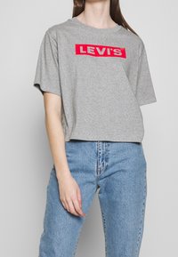Camiseta gris de manga corta con un audaz logo rojo de "LEVI'S" en el pecho. Está hecha de una tela suave, cuenta con un corte relajado y cuello redondo.