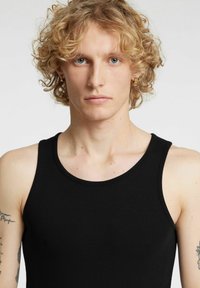 Czarny prążkowany top na szerokich ramiączkach. Posiada okrągły dekolt i dopasowany krój. Modelka ma jasne, kręcone włosy i widoczne tatuaże.