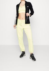 Svart zip-up jacka, neongul crop top och matchande sweatpants med elastiska manschetter. Svarta sneakers med vita detaljer.