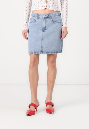 Femme portant une jupe en jean bleu clair, un chemisier court blanc avec un petit imprimé floral rouge, et des mules rouges à talons avec des embouts métalliques.