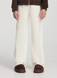 Pantaloni bianchi a gamba larga in materiale morbido, caratterizzati da una texture liscia. Scarpe marroni spesse completano il look. Lo sfondo è semplice.