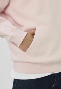 Sweat à capuche rose en matière douce avec une poche avant et des poignets côtelés. Superposé à une chemise blanche, associé à un jean foncé.