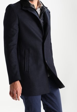 Man wearing a dark navy overcoat with side pockets over a patterned shirt and navy pants, standing against a plain light background.