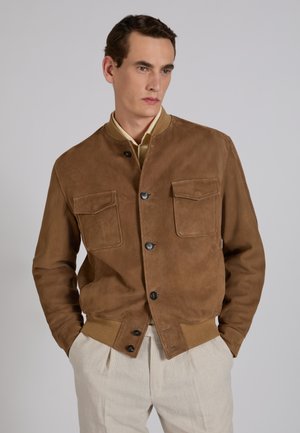 Mann trägt eine braune Wildlederjacke mit Knopfleiste und Klappentaschen, kombiniert mit hellbeigen Hosen, steht mit den Händen in den Taschen.