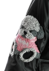 Chaqueta negra con un parche de perro texturizado y peludo en la manga en tonos grises y rosas, con detalles bordados y acentos negros contrastantes.