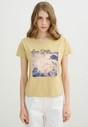 Joven con cabello rubio que lleva una camiseta amarilla con estampado floral y el texto "No pidas permiso para florecer."