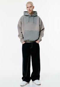 Hættetrøje i gradient grå med frontlomme og synlig syning, parret med sorte vidde bukser og hvide sneakers.