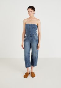 Denimsoverall med strapless topp, vida avklippta ben och frampocketar. Bärs med bruna mocka slip-on skor. Ljusblå färg.