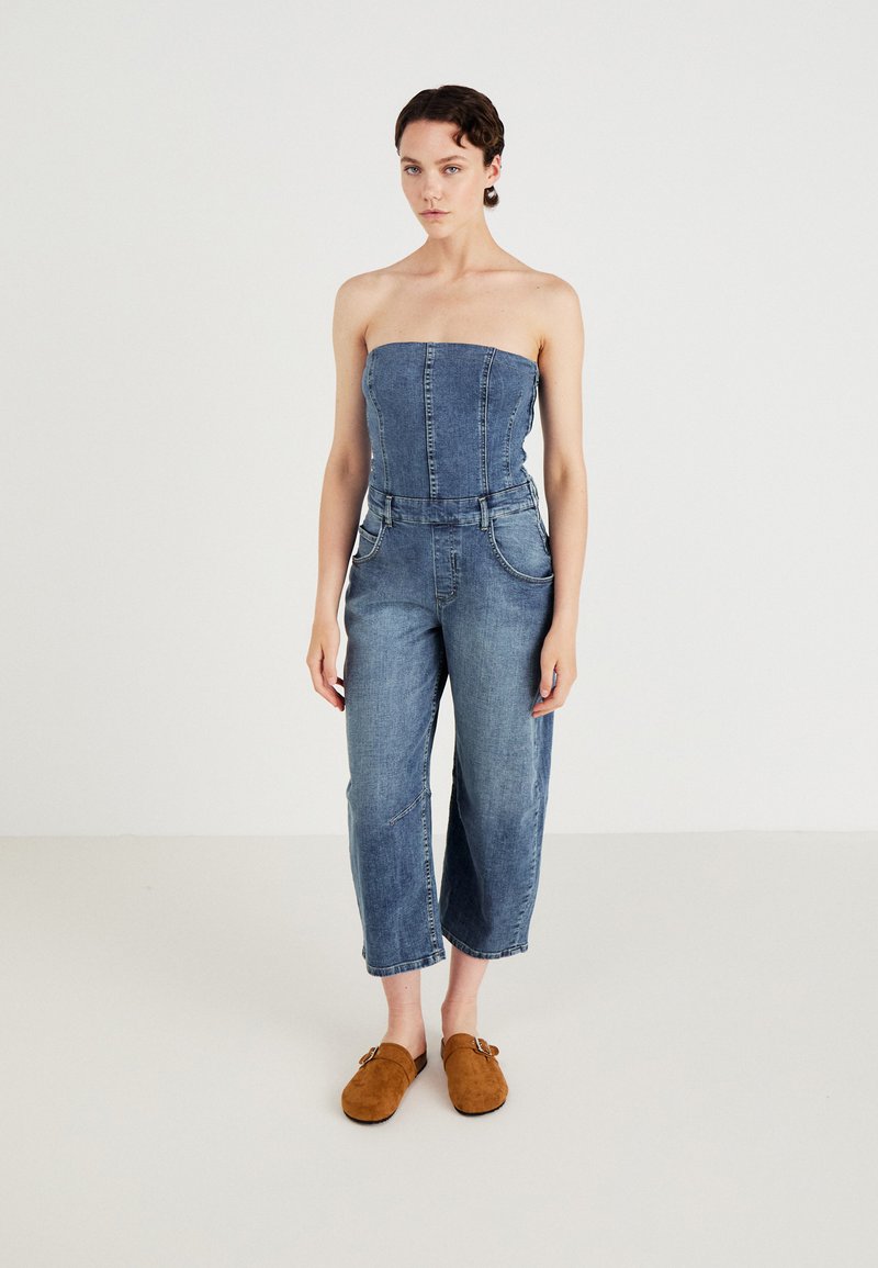Denimsoverall med strapless topp, vida avklippta ben och frampocketar. Bärs med bruna mocka slip-on skor. Ljusblå färg.