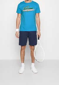 Turkos t-shirt med en grön och vit logotyp, i kombination med marinblå shorts och vita sneakers. Håller en vit tennisracket.