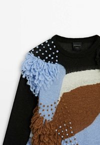 Mehrfarbiger gestrickter Pullover mit schwarzen, hellblauen, braunen und cremefarbenen Abschnitten; zeichnet sich durch strukturierte Wolle, flauschige Akzente und Punkte aus.