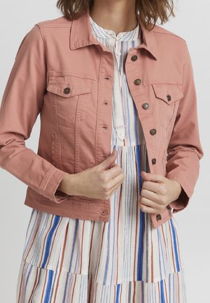 Femme ajustant une veste rose poudré boutonnée sur une robe rayée verticale multicolore avec des tons bleus, marron, noir et blanc.