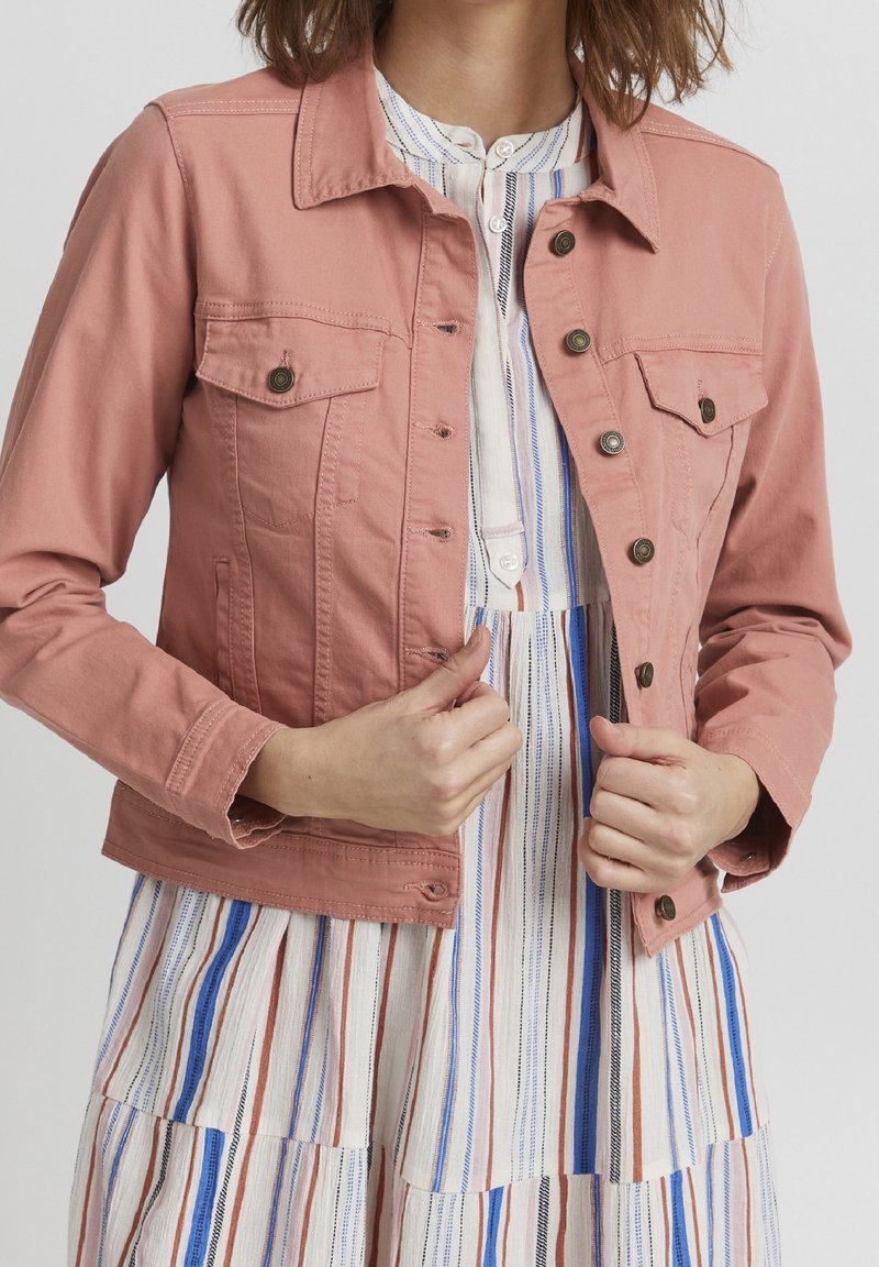 Femme ajustant une veste rose poudré boutonnée sur une robe rayée verticale multicolore avec des tons bleus, marron, noir et blanc.