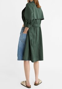 Zielony płaszcz trench o talii związanej w pasie, nałożony na dżinsową spódnicę. Płaszcz ma szeroki kołnierz i podwinięte rękawy, a całość uzupełniają beżowe obuwie.