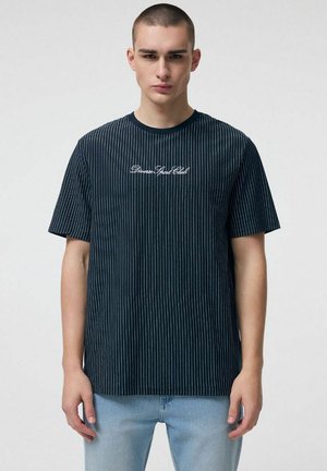 Młody mężczyzna o krótkich włosach, ubrany w czarną koszulkę w białe prążki z białym napisem "Diverse Sport Club" oraz jasnoniebieskie dżinsy, stojący na jednolitym tle.