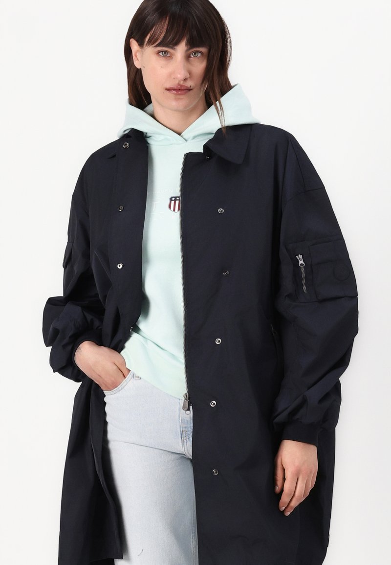 Abrigo largo marino con un corte relajado, botones a presión y un bolsillo con cremallera. Lleva un hoodie ligero color menta y unos jeans de color azul claro.