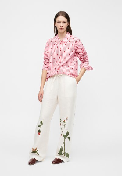 Woman wearing a pink and white striped shirt with small red fruit prints and white wide-leg pants with botanical and bee embroidery.