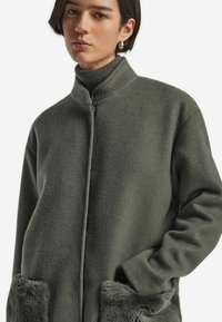 Olivgrön fleecejacka med hög krage, framdragkedja och mjuka fickor med pälsfoder. Har en avslappnad passform och slät textur.