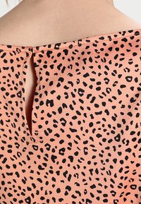 Tejido color durazno con manchas irregulares de leopardo negras, que presenta una pequeña abertura vertical y detalle de costura cerca del escote sobre la piel.