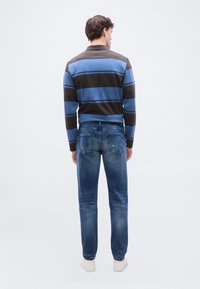 Άνδρας που στέκεται με την πλάτη προς τα εμπρός, φορώντας μπλε και καφέ ριγέ μακρυμάνικο πουκάμισο, ξεθωριασμένο μπλε τζιν και λευκά αθλητικά παπούτσια σε απλό φόντο.