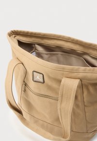 Sac fourre-tout en toile beige avec deux poignées, poche zippée à l'avant, compartiment principal ouvert et petit patch logo carré sur le devant.