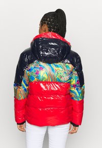 Veste rembourrée avec un corps rouge brillant, des manches noires et un design marbré multicolore dans le dos. Elle possède une capuche amovible.