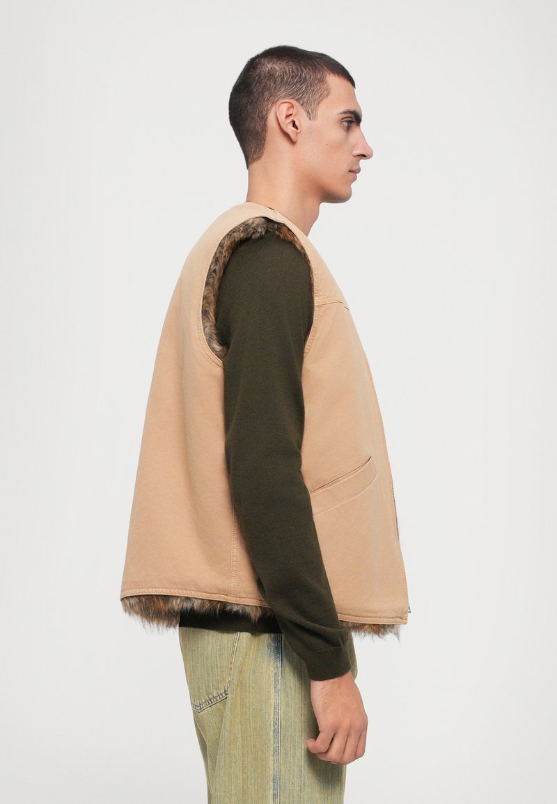Tan sleeveless vest with a soft fur lining, featuring a single chest pocket, worn over a dark green long-sleeve shirt and light denim pants.