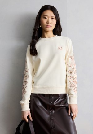 Νεαρή γυναίκα που φοράει ένα κρεμ πουλόβερ με κεντημένα μανίκια και ένα σκούρο καφέ δερμάτινο φούστα με κουμπιά, κρατώντας μια μαύρη τσάντα στο πλάι της.