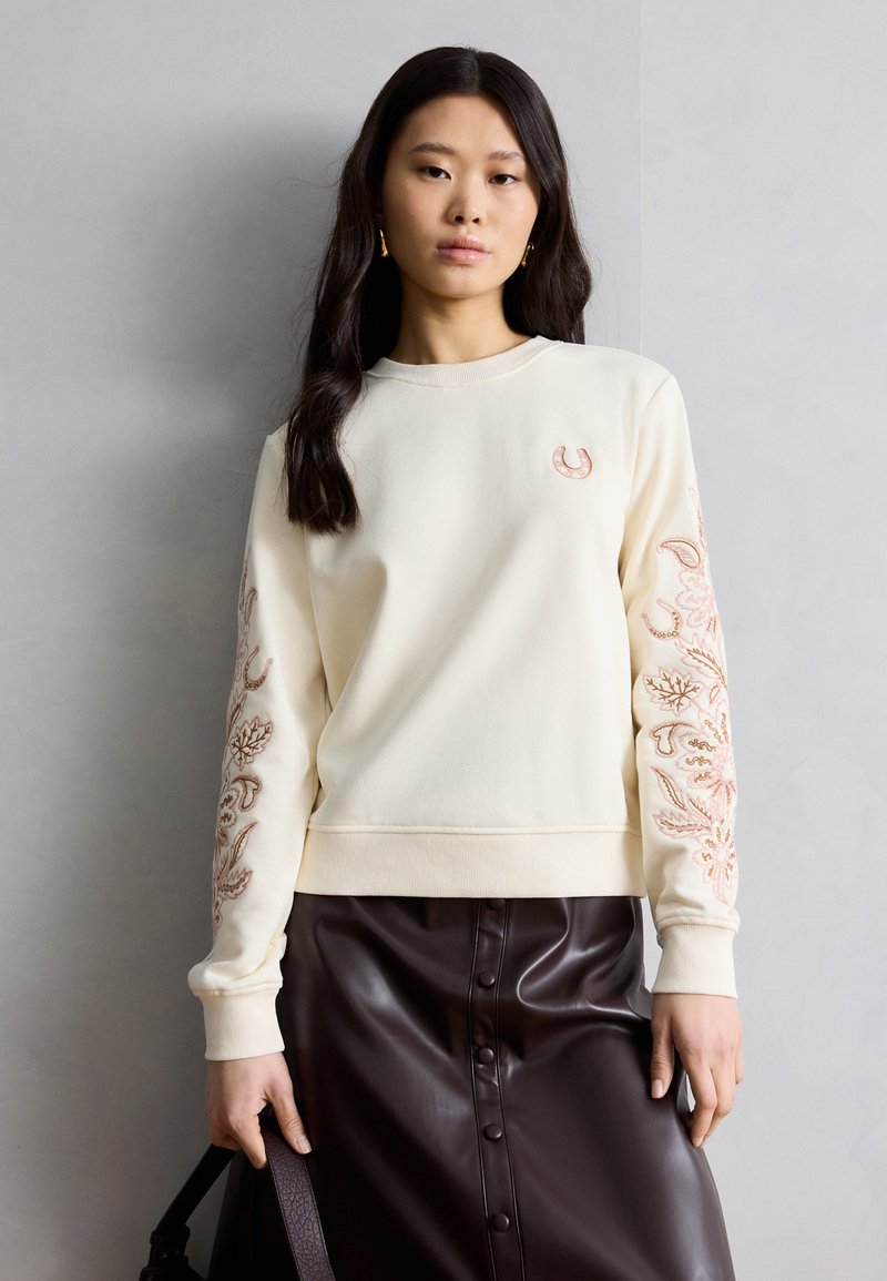 Νεαρή γυναίκα που φοράει ένα κρεμ πουλόβερ με κεντημένα μανίκια και ένα σκούρο καφέ δερμάτινο φούστα με κουμπιά, κρατώντας μια μαύρη τσάντα στο πλάι της.