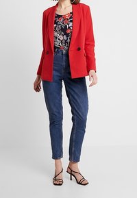 Blazer rosso con rever a incavo e due bottoni, abbinato a una camicetta floreale, jeans blu a vita alta e tacchi neri con cinturini.