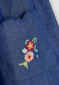 Denim tkanina ima zakovanu džepu s valovitim rubom i višebojnim cvjetnim vezom, uključujući crvene, žute, ružičaste i plave cvjetove na plavoj pozadini.