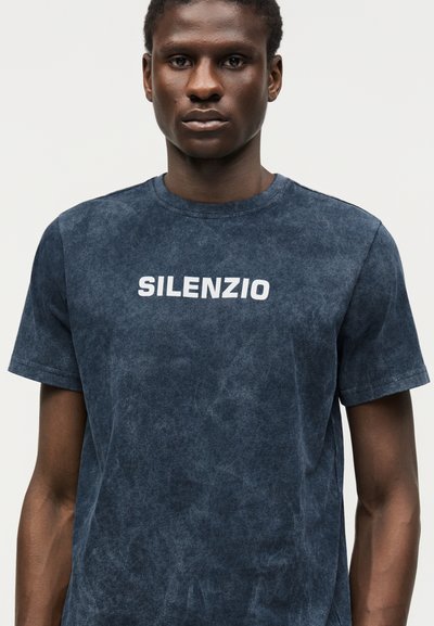 Άνδρας φοράει σκούρο μπλε, ραβδωτό T-shirt με τη λέξη "SILENZIO" τυπωμένη με λευκά γράμματα στο στήθος, με απλό φόντο.