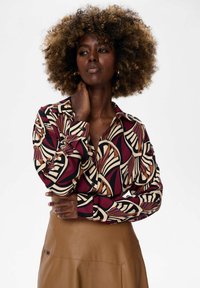 Woman with curly hair wears a patterned long-sleeve blouse in burgundy, cream, and black, and a tan leather skirt, posing with hand on neck.