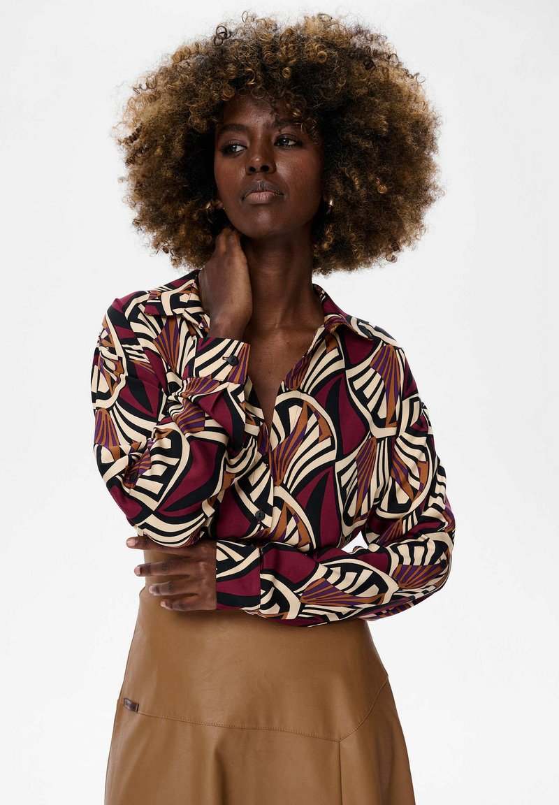 Woman with curly hair wears a patterned long-sleeve blouse in burgundy, cream, and black, and a tan leather skirt, posing with hand on neck.