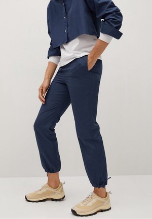Personne portant un pantalon de jogging bleu marine avec des poignets élastiques, une chemise blanche sous une veste courte bleu marine, et des chaussures de sport beiges.