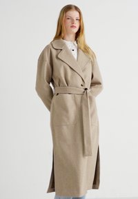 Trench coat de mezcla de lana beige con cintura ceñida, cuello de solapa, bolsillos laterales y una silueta larga con aberturas laterales.