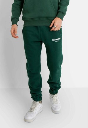 Groene katoenen joggers met ribgebreide boorden en zakken, met een witte "SIXTHJUNE PARIS" logo aan de linkerkant, gecombineerd met witte sneakers.