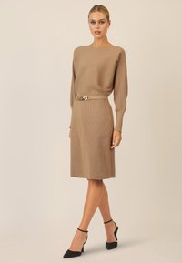 Strickkleid in Beige mit gerippter Textur, ausgestattet mit einem Bootsausschnitt, langen Ärmeln und einem Gürtel mit einem goldenen Kettenakzent. Schwarze High Heels runden das Outfit ab.
