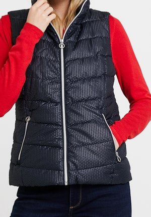 Žena s černou texturovanou prošívanou vestou přes červenou košili s dlouhým rukávem, jednou rukou v kapse a druhou u límce.